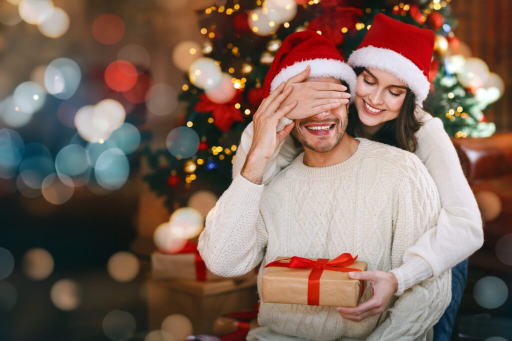 Jeune femme offrant un cadeau surprise à son mari, qui ferme les yeux, célébrant Noël à la maison.