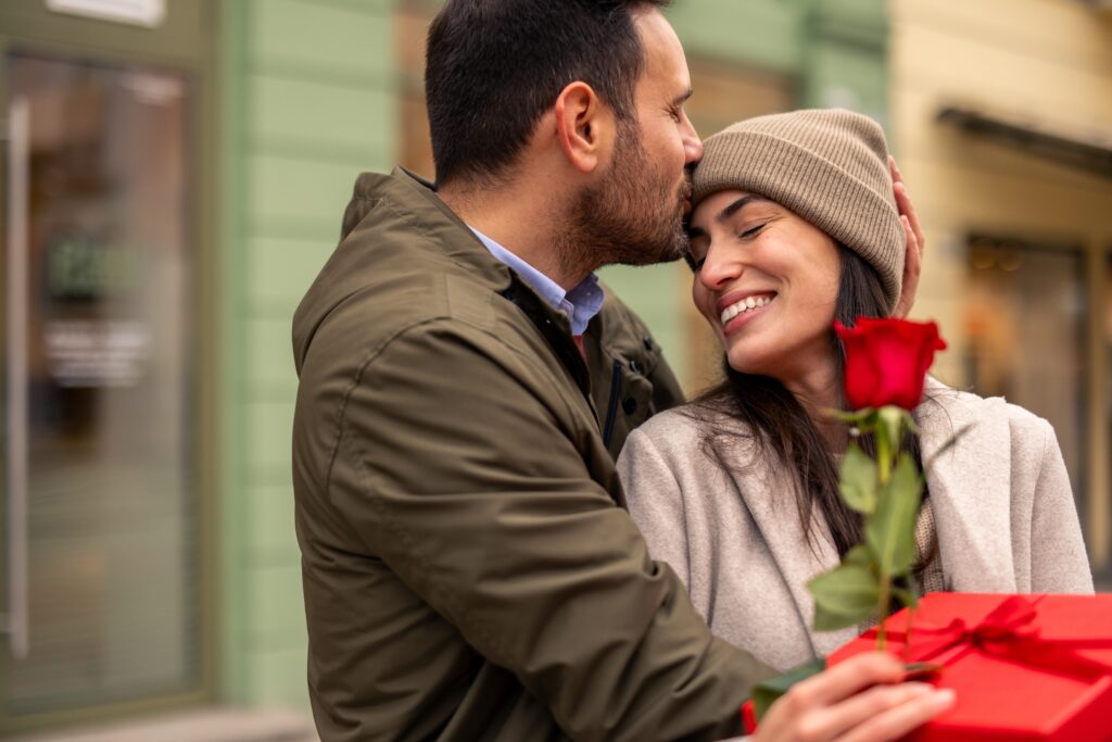 Un couple s'offrent des cadeaux à l'occasion de la Saint-Valentin. L'homme porte une veste verte et serre dans ses bras sa partenaire, qui porte un manteau beige. Ils tiennent une rose rouge et font leur cadeau en plein air, dans un environnement urbain.