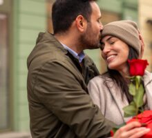 Un couple s'offrent des cadeaux à l'occasion de la Saint-Valentin. L'homme porte une veste verte et serre dans ses bras sa partenaire, qui porte un manteau beige. Ils tiennent une rose rouge et font leur cadeau en plein air, dans un environnement urbain.