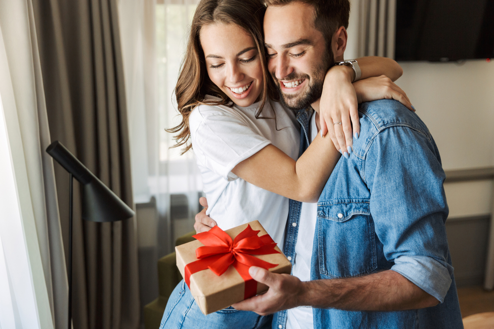 Beau jeune couple amoureux à la maison, célébrant la saint-valentin l'échange d'un paquet cadeau, s'embrassant.