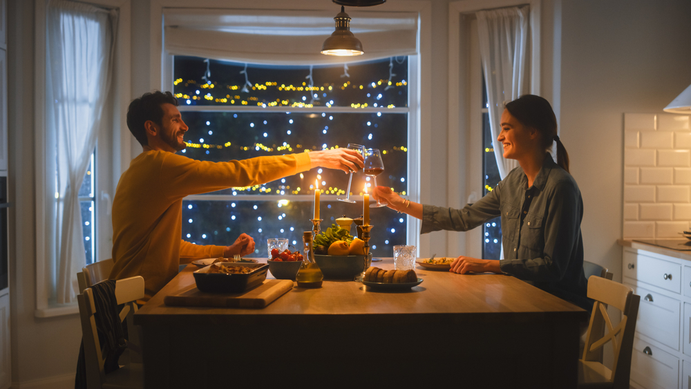 Un jeune couple heureux et amoureux prend un dîner romantique pour la saint-valentin, trinque avec des verres de vin, mange un repas savoureux dans la cuisine, et discute.