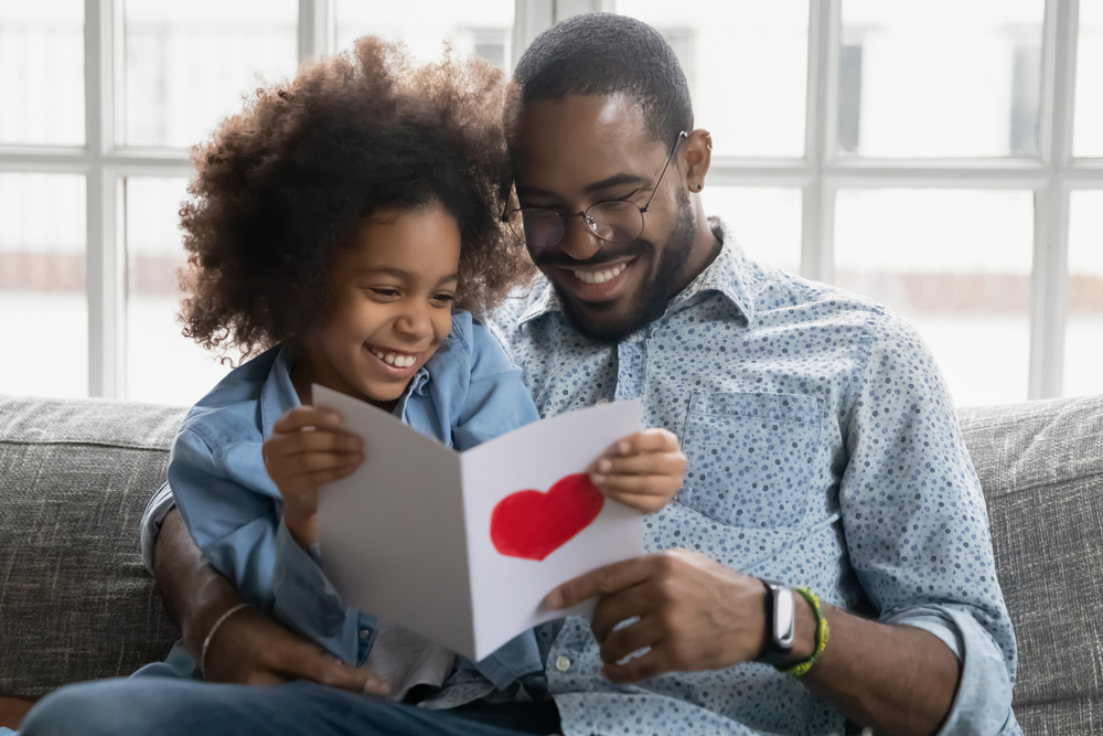 La petite fille a fait une surprise à son père, assis sur le canapé  en lisant des mots agréables et chaleureux sur une carte qu'elle a fait pour la Fête des Pères, avec un grand coeur rouge en signe d'amour pour son père.