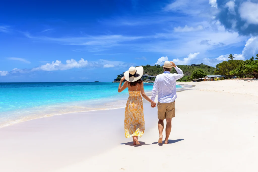 Un couple en lune de miel, coiffé de chapeaux de soleil, se promène sur une plage tropicale bordée par une mer turquoise dans les Caraïbes, sur l'île d'Antigua.
