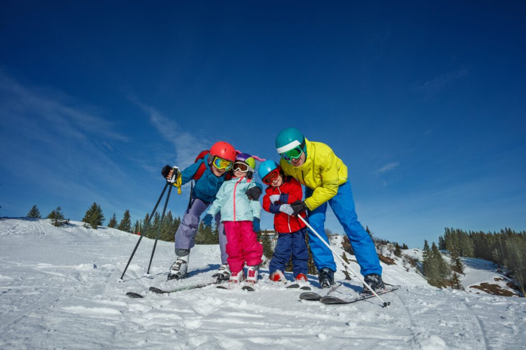 Une famille de quatre personnes, vêtue d'un équipement de ski coloré, de casques, pose joyeusement ensemble et se serre sur une pente enneigée sous un ciel bleu clair