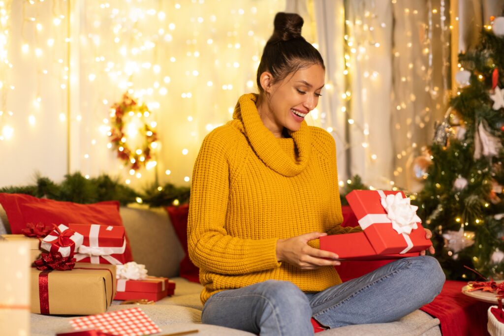 Femme excitée ouvrant un coffret Smartbox reçue pour Noël assis sur le canapé à la maison, dans une salle décorée avec des guirlandes lumineuses festives.