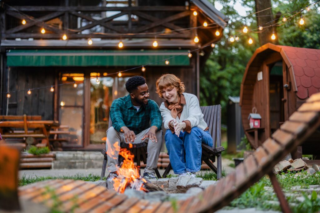 Un couple se détend au coin d'un feu de camp chaleureux dans un cadre rustique extérieur, partageant une conversation joyeuse sous les lumières de la maison.