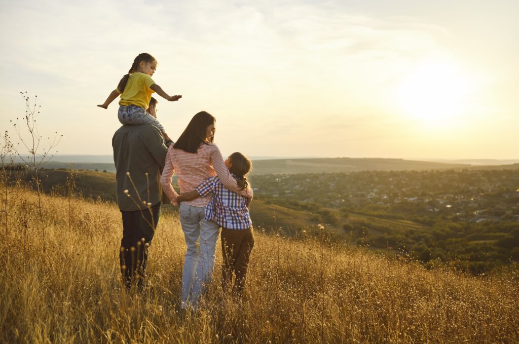 Une famille en week-end pour la fête des pères au coucher du soleil, les parents serrent les enfants à l'extérieur. La lumière dorée réchauffe les prés alors que la nature les entoure, surplombant les collines, partageant des sourires dans un moment calme.