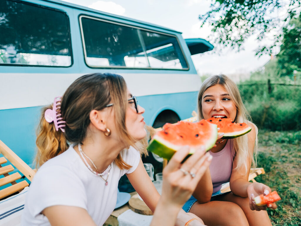 Dos amigas sentadas delante de una caravana riendo y comiendo sandía